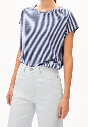 Kvinde iført blå og hvide horisontale stribede kortærmede top, stoppet ned i højttaljede lyseblå jeans, mod hvid baggrund.