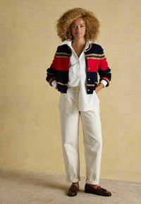 Marineblau-rote und grüne gestreifte Strickjacke über einem weißen Hemd, kombiniert mit beige Hosen und braunen Mokkasins. Modell steht mit Händen in den Taschen.