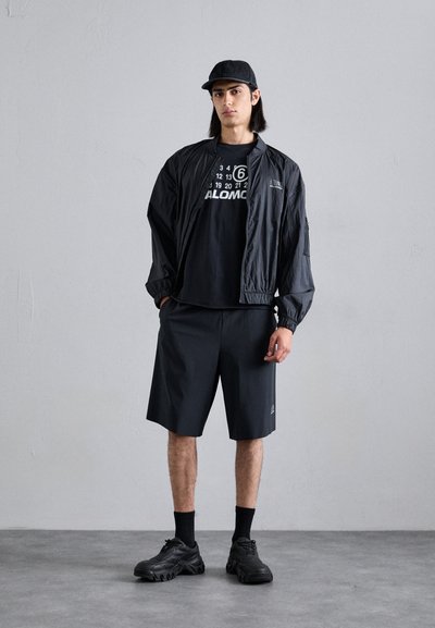 Veste légère noire à coupe décontractée sur un t-shirt graphique noir, associée à un short noir, des chaussettes noires et des sneakers noires chunky.