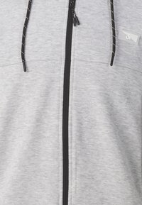 Felpa grigia chiara con zip, cerniera nera e coulisse nera, con punti bianchi e una piccola patch logo trasparente sul petto.