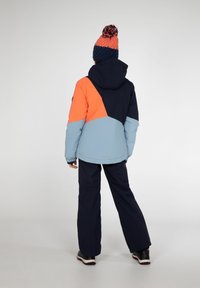 Farbblockierte Jacke mit orangefarbenen, marineblauen und hellblauen Einsätzen, ausgestattet mit einer Kapuze und verstellbaren Bündchen, kombiniert mit marineblauen Schneehosen und Stiefeln.