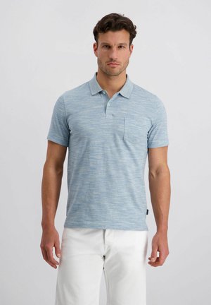 Mann steht, trägt ein hellblaues Poloshirt mit kurzen Ärmeln und Brusttasche sowie weiße Hose vor einem einfarbigen weißen Hintergrund.