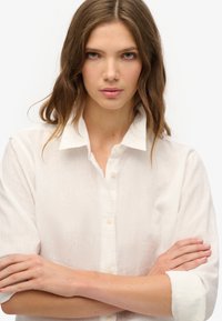 Biała lniana koszula z guzikami z przodu, z zagiętymi rękawami i wykładanym kołnierzykiem. materiał wydaje się lekki z subtelną teksturą.