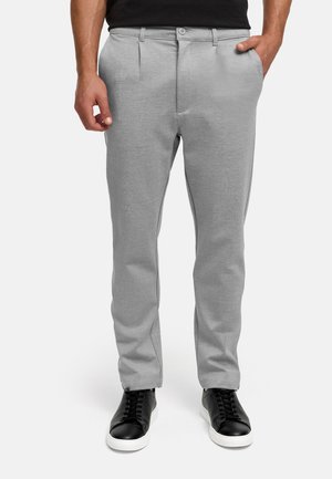 Mann trägt hellgraue Slim-Fit-Hose mit Seitentaschen und schwarze Low-Top-Sneaker mit weißen Sohlen, stehend vor weißem Hintergrund.
