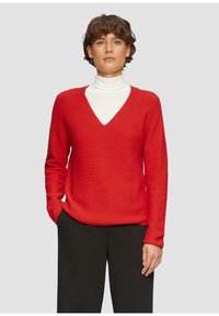 Rode gebreide trui met een V-hals en geribbeld textuur, gecombineerd met een witte coltrui. Het model draagt zwarte broek, met handen in de zakken.