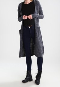 Lång grå cardigan med texturerad stickning, öppen framsida och två sidofickor, bärs över en svart topp och kombineras med mörka skinny jeans och svarta stövlar.