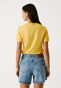 Gelbes Baumwoll-T-Shirt mit kurzen Ärmeln; getragen mit hochtaillierten blauen Denim-Shorts mit fransigem Saum und gestickten Mustern auf den Gesäßtaschen.