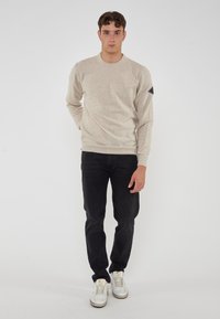 Giovane uomo che indossa un maglione beige, jeans neri e sneaker bianche, in piedi contro uno sfondo bianco uniforme.