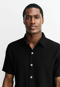 Sort kortærmet skjorte med ribbet tekstur, der har en krave og hvide knapper. Stofdet ser blødt og let ud.