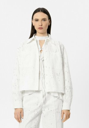 Frau mit glattem, dunklem Haar, die eine weiße, strukturierte Jacke, eine Rüschenbluse mit Schlüssellochausschnitt und passende weiße Hosen trägt, in neutraler Haltung stehend.