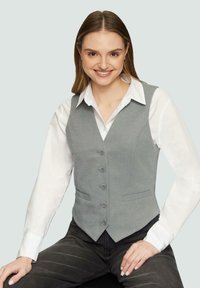 Grijze op maat gemaakte vest met een v-hals, voorzijde met knoopsluiting, twee zijzakken, gedragen over een witte blouse met kraag, gecombineerd met donkere spijkerbroeken.