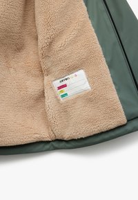 Grøn yderjakke med en beige, fluffy fleece inderside. Har et mærke til plejeinstruktioner fastgjort til foret.