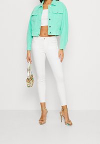 Turkusowa dżinsowa kurtka, dopasowany biały top cropped, białe skinny jeans, wielokolorowa torebka oraz sandały na obcasie z paskami i złotymi akcentami.