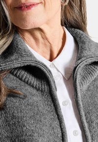 Pull en maille grise avec un col zippé, porté sur une chemise blanche à col avec des boutons ; met en avant les détails de tissu lisse et côtelé.