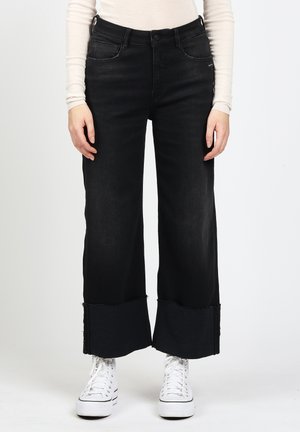 Frau trägt schwarze Jeans mit weitem Bein und rohem Saum sowie weiße High-Top-Sneaker, steht vor einem schlichten weißen Hintergrund.
