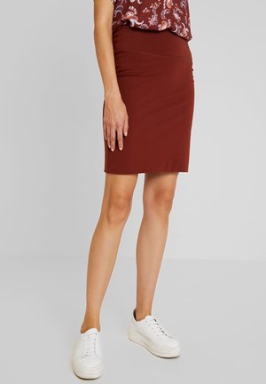 Femme portant une jupe marron ajustée jusqu'au genou, un chemisier à motifs et des baskets à plateforme blanches, debout devant un fond uni gris clair.