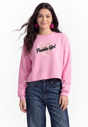 Ung kvinde med langt mørkt hår iført pink sweatshirt med hjerte og teksten "Pookie Girl", stjerneformede øreringe samt blå jeans foran hvid baggrund.