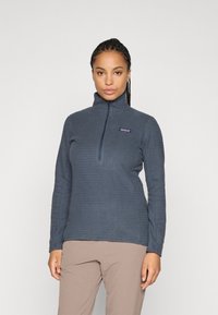 Vrouw in een blauw gestructureerd fleecejack met kwartrits en lichtbruine broek, staande tegen een effen lichtgrijze achtergrond.