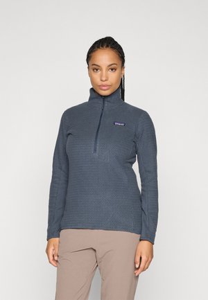 Vrouw in een blauw gestructureerd fleecejack met kwartrits en lichtbruine broek, staande tegen een effen lichtgrijze achtergrond.
