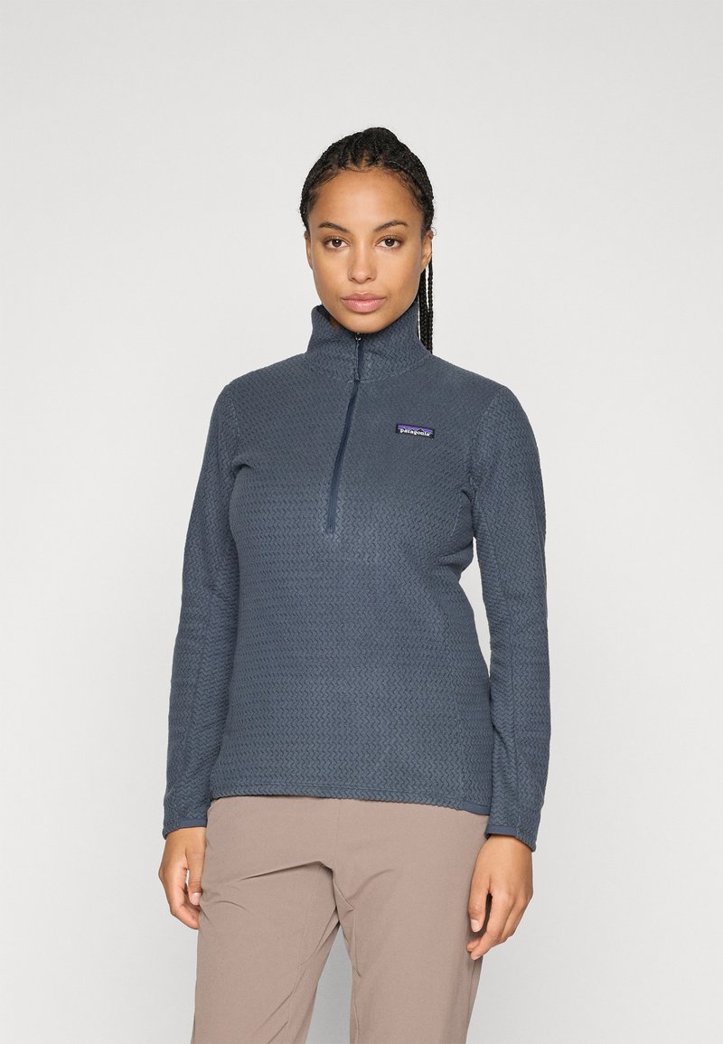 Vrouw in een blauw gestructureerd fleecejack met kwartrits en lichtbruine broek, staande tegen een effen lichtgrijze achtergrond.