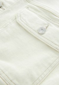 Lyse creme denimstof med en struktureret overflade, der har en sølvfarvet trykknap og en skrå lomme med rene syningsdetaljer.