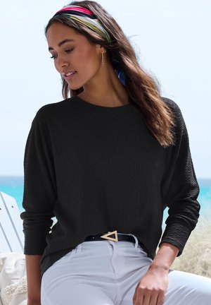 Schwarzes, geripptes Langarmshirt kombiniert mit weißen Hosen, versehen mit einer dreieckigen goldenen Gürtelschnalle und einem farbenfrohen gestreiften Haarband.