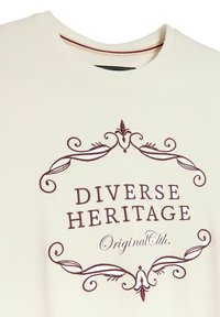 Koszulka w kolorze kremowym z bordowymi ornamentami w kształcie wirów otaczającymi napis "Diverse Heritage Original Clth." wyszyty bordową nicią.