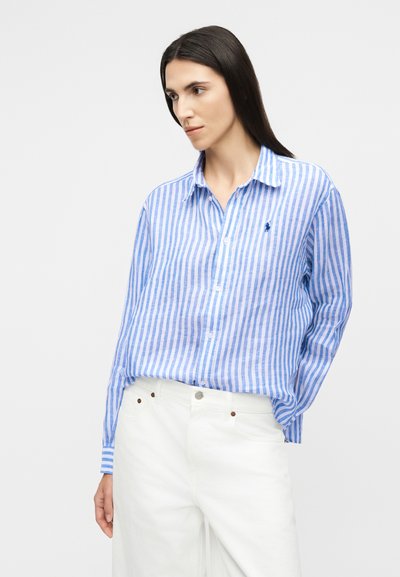 Femme portant une chemise à rayures verticales bleues et blanches rentrée dans un pantalon blanc taille haute, debout devant un fond uni.