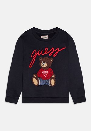 Sweatshirt noir à manches longues avec le texte rouge « guess » et un ours en peluche brun souriant portant un pull rouge Guess et un pantalon bleu à l'avant.