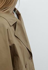 Hellbrauner Trenchcoat mit großem Kragen, einreihigem Design und auffälligen Knöpfen. Der Stoff wirkt glatt mit einer leicht strukturierten Oberfläche.