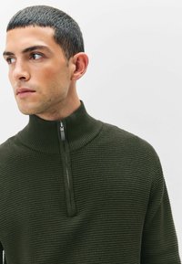 Pulover texturat de culoare verde închis, cu guler înalt, având închidere prin fermoar în față și detalii ribbate la guler și la manșete.