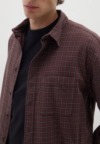 Camicia flanella a quadri marrone con colletto button-down, dotata di taschino e linee a griglia bianche in contrasto su una t-shirt nera sottostante.
