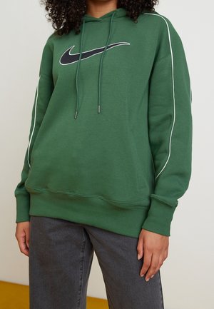 Personne portant un sweat à capuche vert Nike avec un logo Swoosh noir, des bandes blanches sur les manches et un jean gris foncé, debout devant un fond uni.