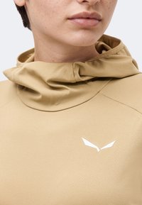 Κοντινό πλάνο ατόμου που φοράει μπεζ ένδυμα με κουκούλα και μικρό λευκό γωνιώδες λογότυπο στο στήθος, που δείχνει λεπτομέρειες του λαιμού και του κάτω μέρους του προσώπου.