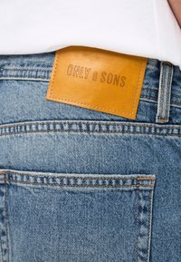 Svijetloplave traperice s smešnim kožnim etiketom utisnutim s natpisom "ONLY & SONS" na stražnjem pojasu. Vidljivi detalji šavova.