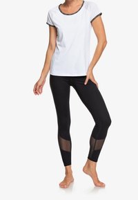 Mujer vistiendo una blusa blanca de manga corta con ribete negro y leggings negros con paneles de malla transparente en la parte baja de las piernas, de pie descalza.