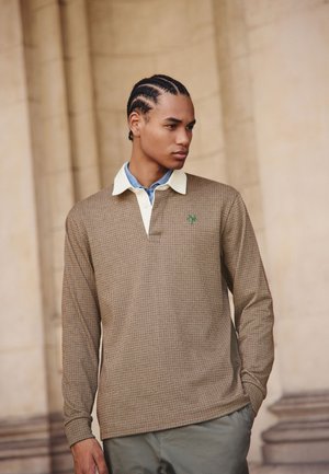 Mlad moški z pletenicami, oblečen v olivno rjavo karo dolgo polo majico s podloženo modro srajco, stoji pred kamnitim zidom.