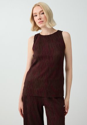 Femme aux cheveux blonds courts portant un haut sans manches texturé marron foncé et un pantalon assorti sur un fond clair uni.