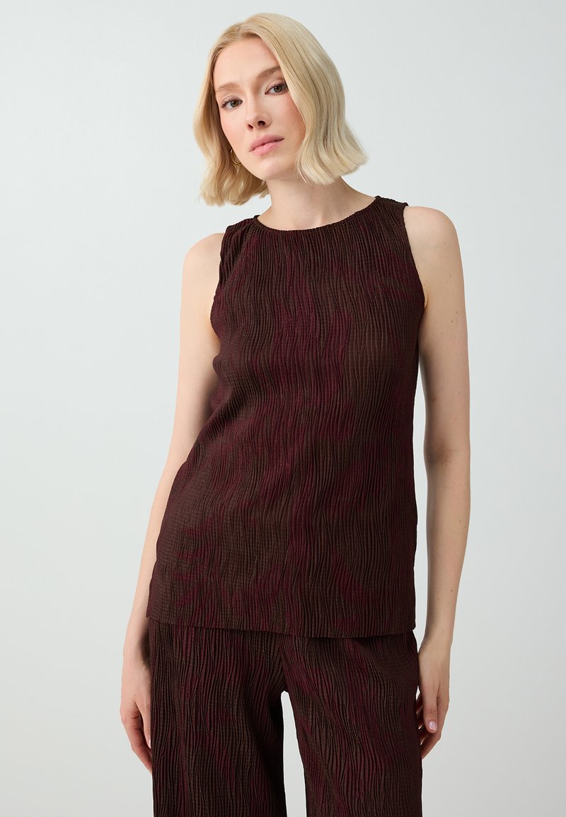 Femme aux cheveux blonds courts portant un haut sans manches texturé marron foncé et un pantalon assorti sur un fond clair uni.