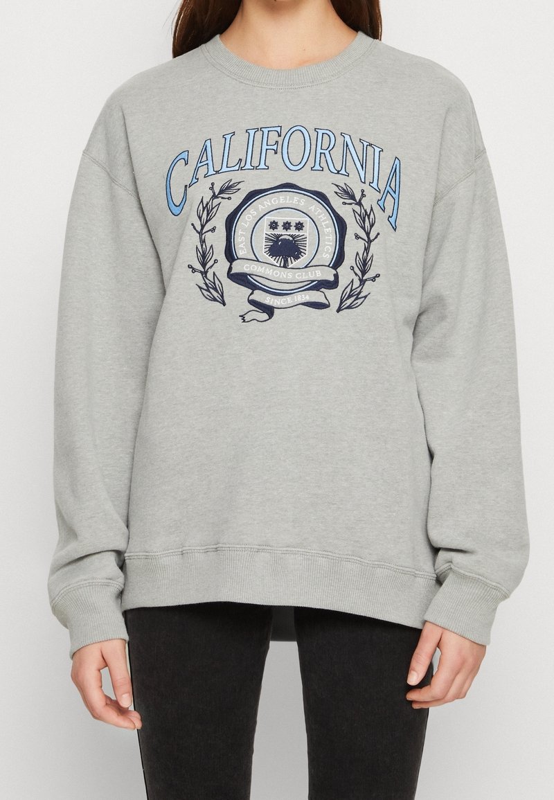 Sudadera gris con texto "CALIFORNIA" en azul. Presenta un emblema gráfico rodeado de ramas de laurel. Corte holgado y puños acanalados.