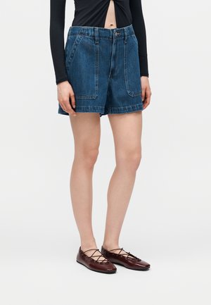 Femeie purtând pantaloni scurți din denim albastru cu talie înaltă, cu buzunare mari în față și balerini maro închis cu șireturi, pe fundal alb.