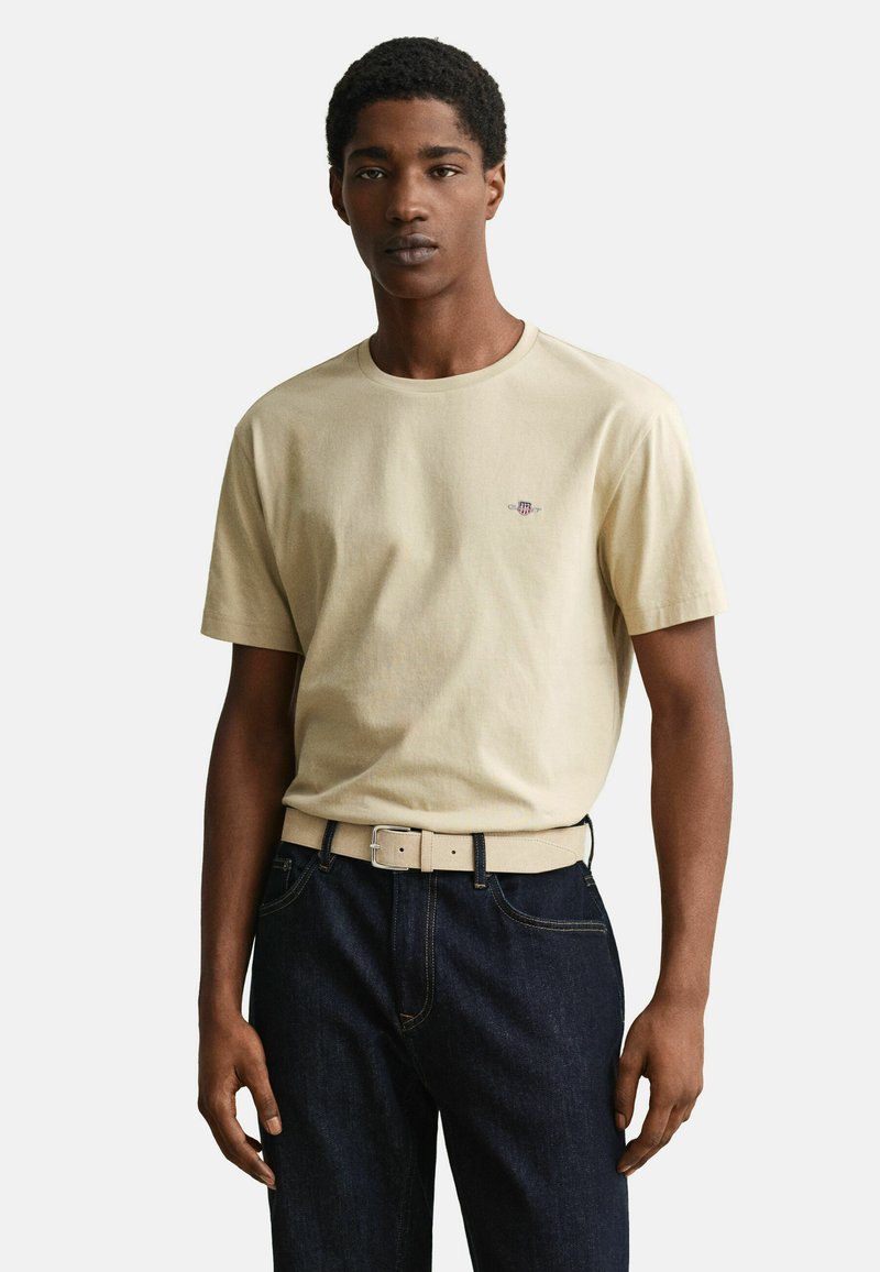 Νεαρός άνδρας που φοράει μπεζ κοντομάνικη μπλούζα με μικρό λογότυπο, σκούρα μπλε τζιν και μπεζ ζώνη, στέκεται μπροστά σε ένα απλό φόντο.