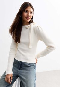 Maglione a coste cream con collo alto e dettaglio a fiocco laterale. Maniche lunghe e silhouette aderente, abbinato a jeans a vita alta.