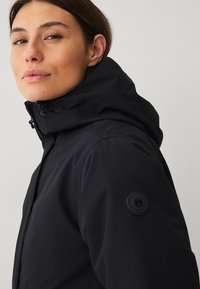 Vrouw in een zwarte hoodie met hoge kraag en ritssluiting aan de voorkant, iets naar links gekeerd tegen een effen lichte achtergrond.