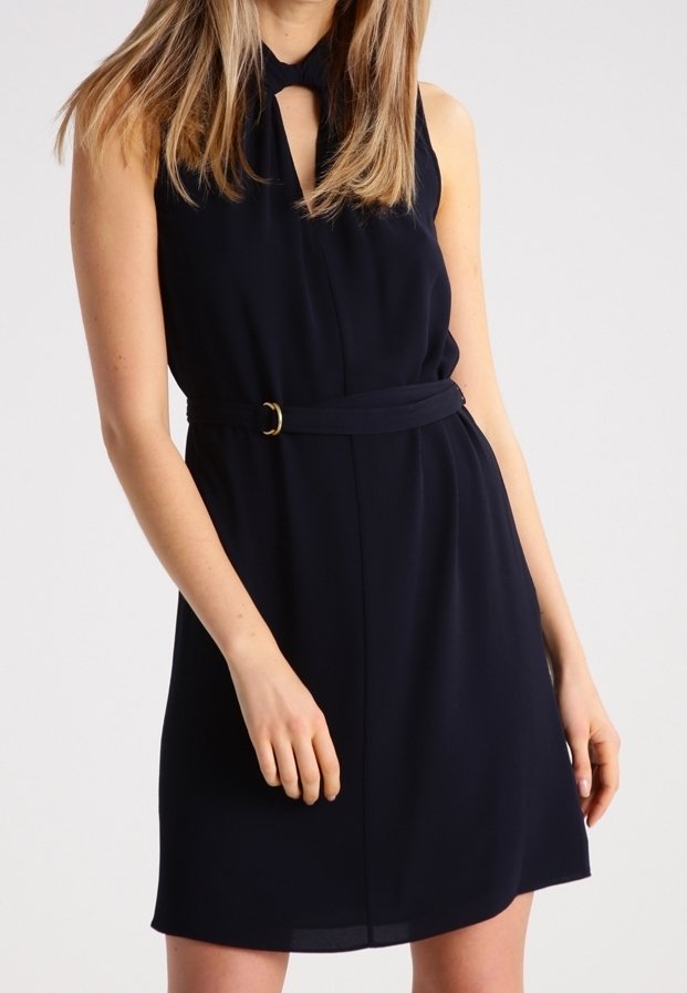Femme portant une robe noire sans manches avec une ceinture à la taille et un décolleté en forme de trou de serrure, debout devant un fond clair uni.