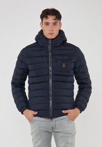 Giacca imbottita blu navy con colletto alto, zip frontale e tasca sul petto con logo. Superficie texturizzata e polsini elasticizzati.