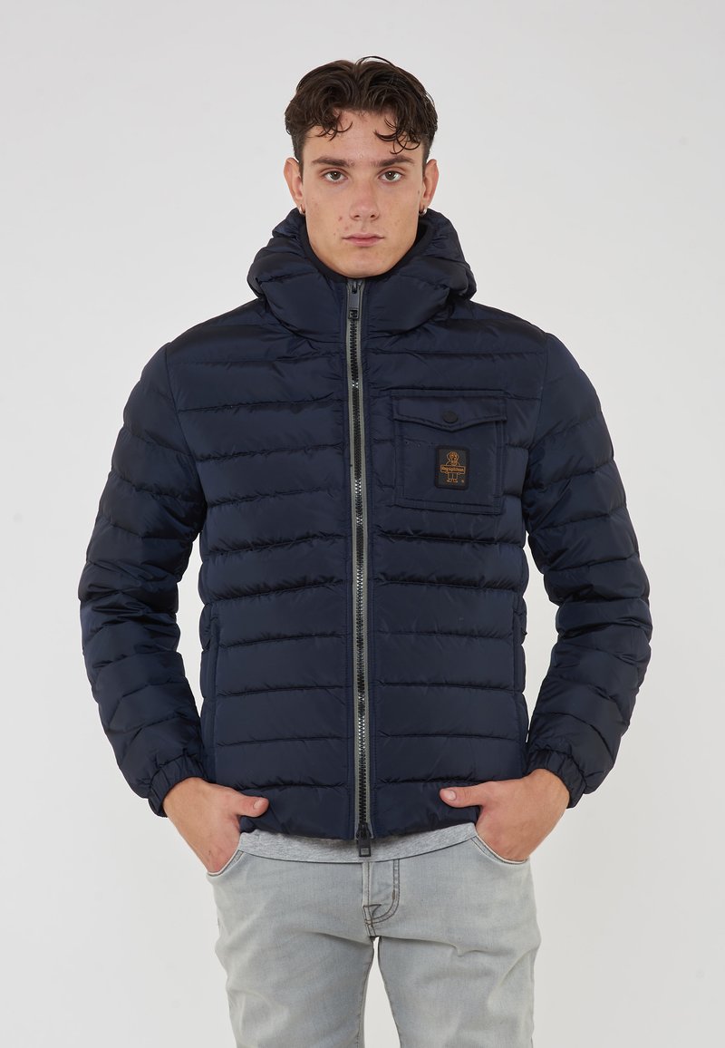Giacca imbottita blu navy con colletto alto, zip frontale e tasca sul petto con logo. Superficie texturizzata e polsini elasticizzati.