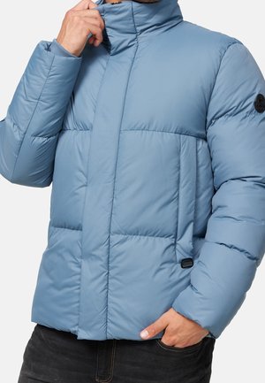 Man wearing light blue quilted puffer jacket with high collar, right hand adjusting collar, left hand in pocket, against plain background.