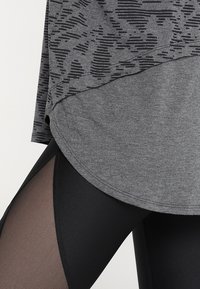 Grå träningstopp med ett texturerat svart mönster ovanför svarta leggings med en genomskinlig nätinsats som lyfter fram en lagerdesign.