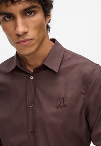Camicia marrone con bottoni, caratterizzata da una texture liscia, con chiusure a bottoni neri e un logo ricamato sul lato sinistro del petto.